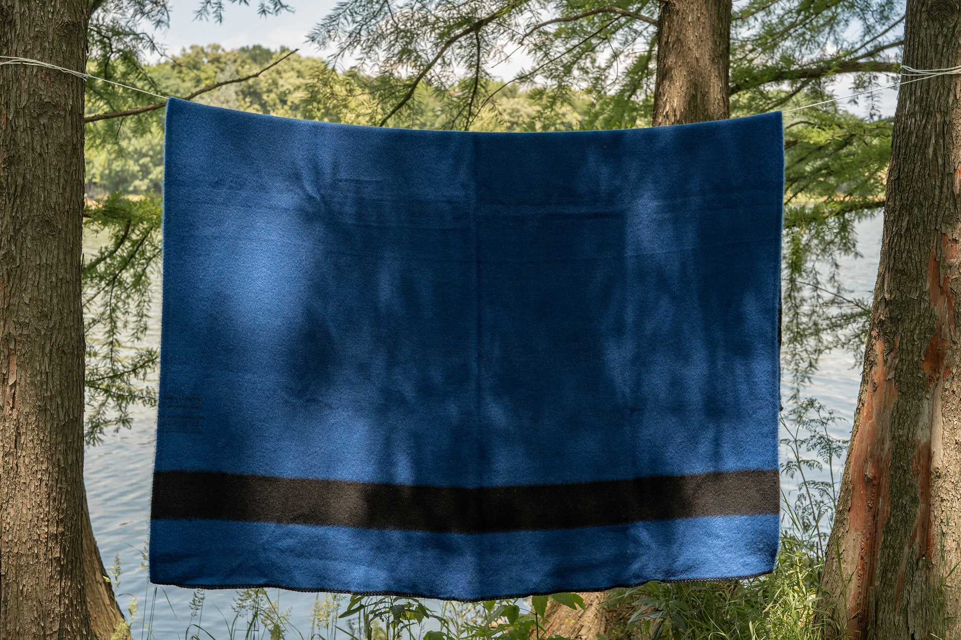 Blue camp woven blanket with a black stripe, made from virgin wool and hanging between two trees by the river.