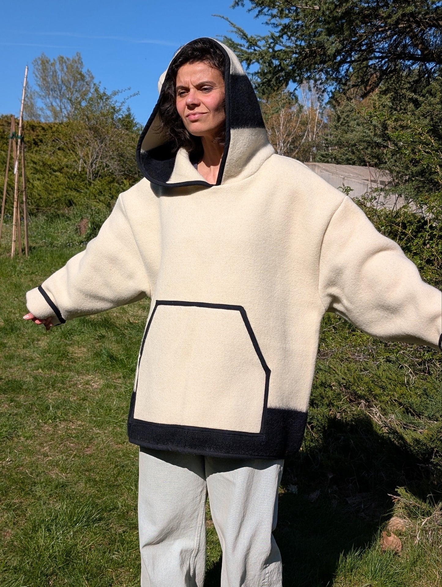 A woman wearing an offwhite wool anorak hoodie with a wide hood and a big front kangaroo pocket. Oversized model suitable for men and women.