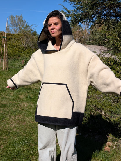 A woman wearing an offwhite wool anorak hoodie with a wide hood and a big front kangaroo pocket. Oversized model suitable for men and women.