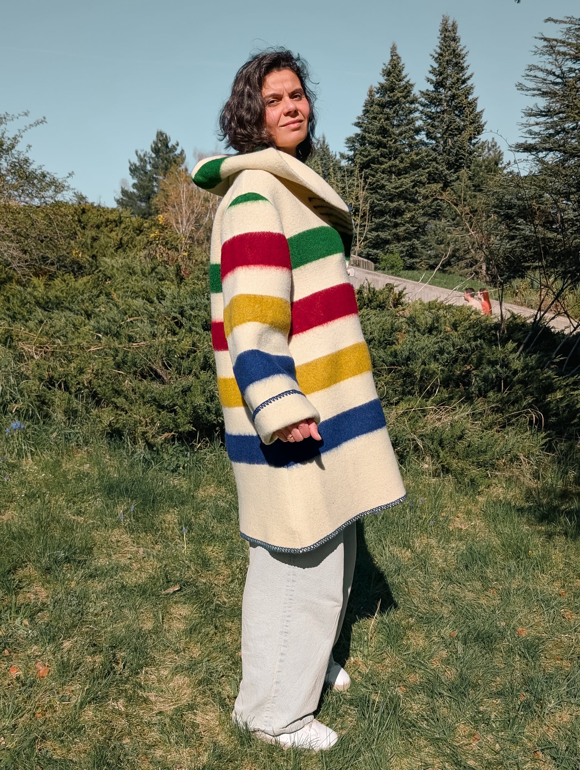 Woman posing with a stylish bespoke upcycled hooded jacket with colorful stripes, outdoors in a park setting.