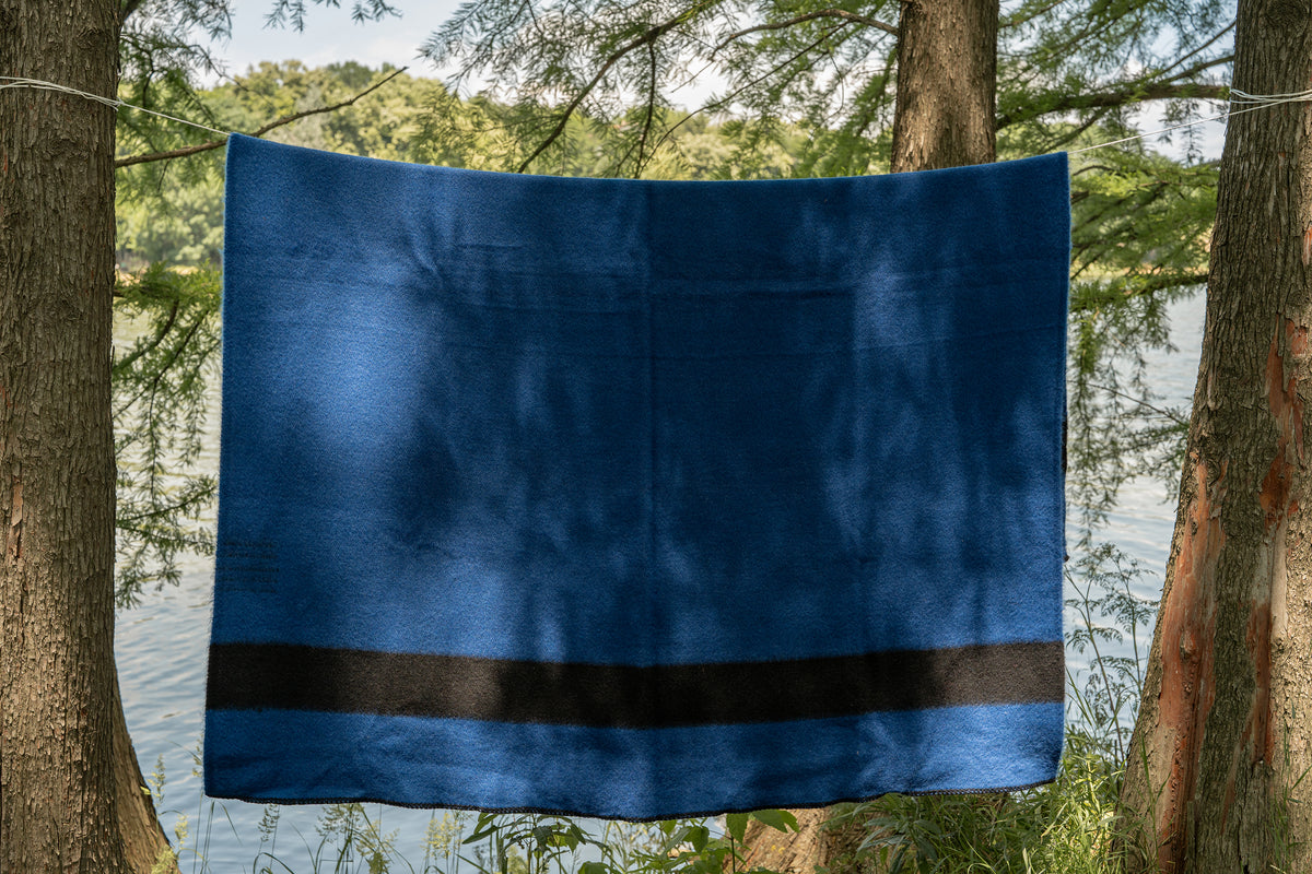 Blue camp woven blanket with a black stripe, made from virgin wool and hanging between two trees by the river.
