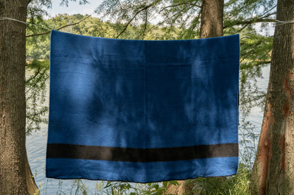 Blue camp woven blanket with a black stripe, made from virgin wool and hanging between two trees by the river.