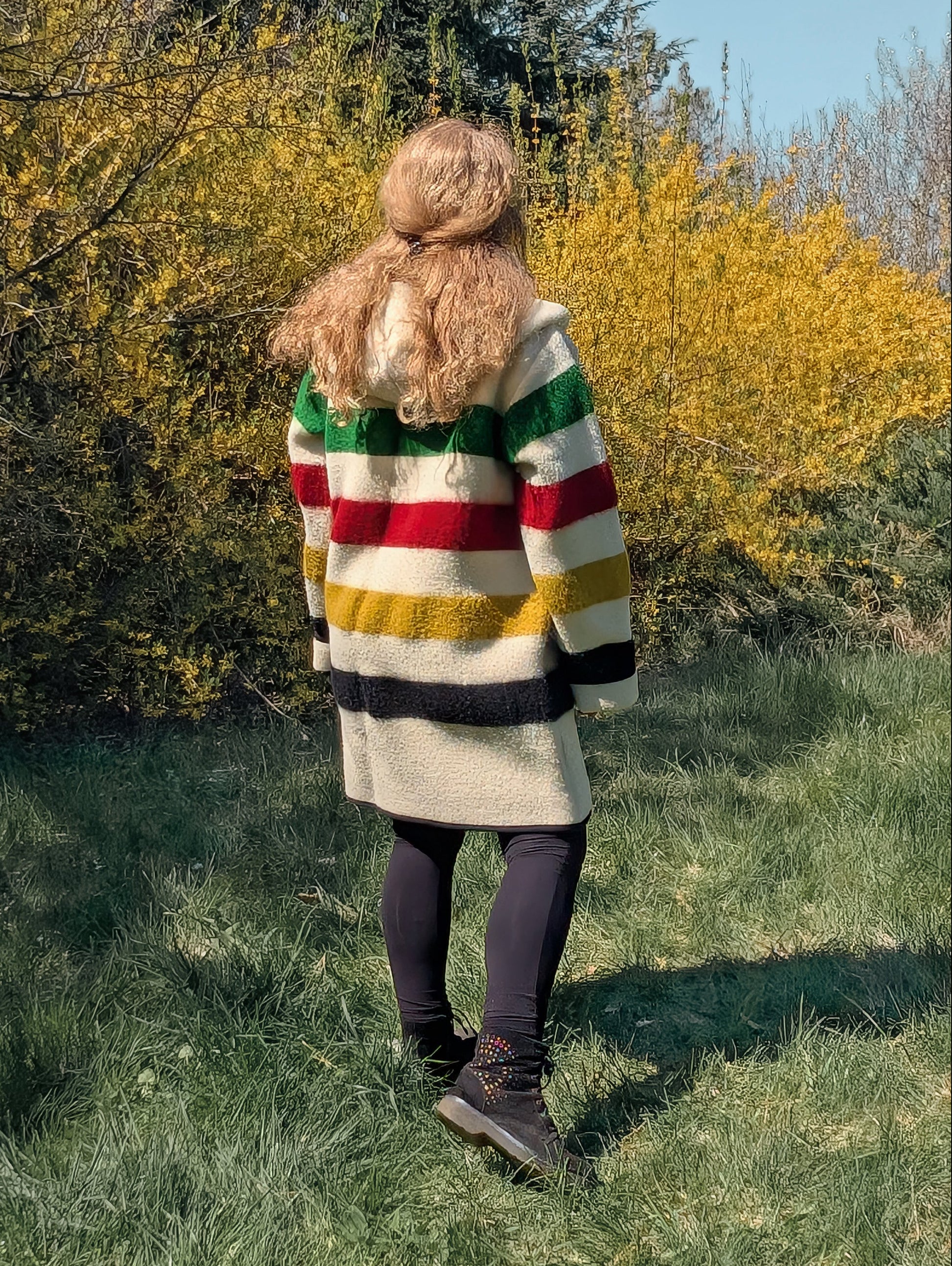 Person wearing a colorful striped coat standing in a grassy area with trees and bushes. 