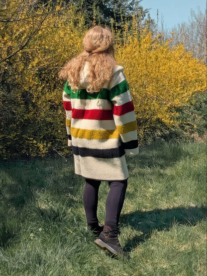 Person wearing a colorful striped coat standing in a grassy area with trees and bushes. 