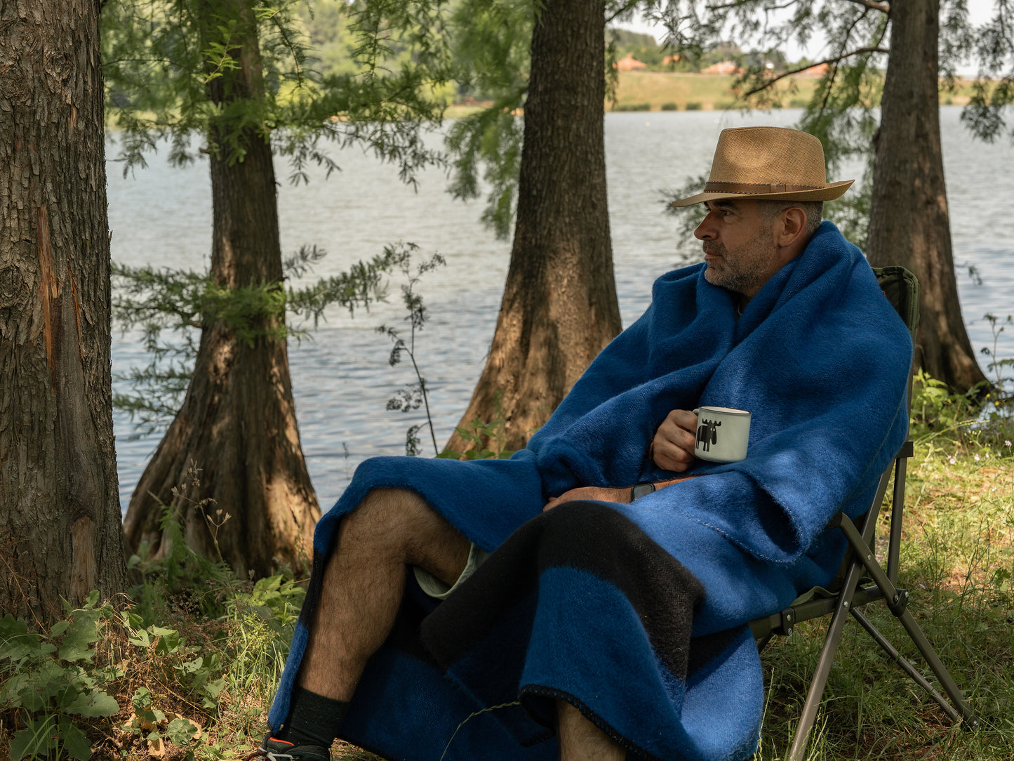 Person wrapped in a blue pioneer blanket with a black stripe, made of natural wool. The man is sitting in a chair by a lake, holding a mug.