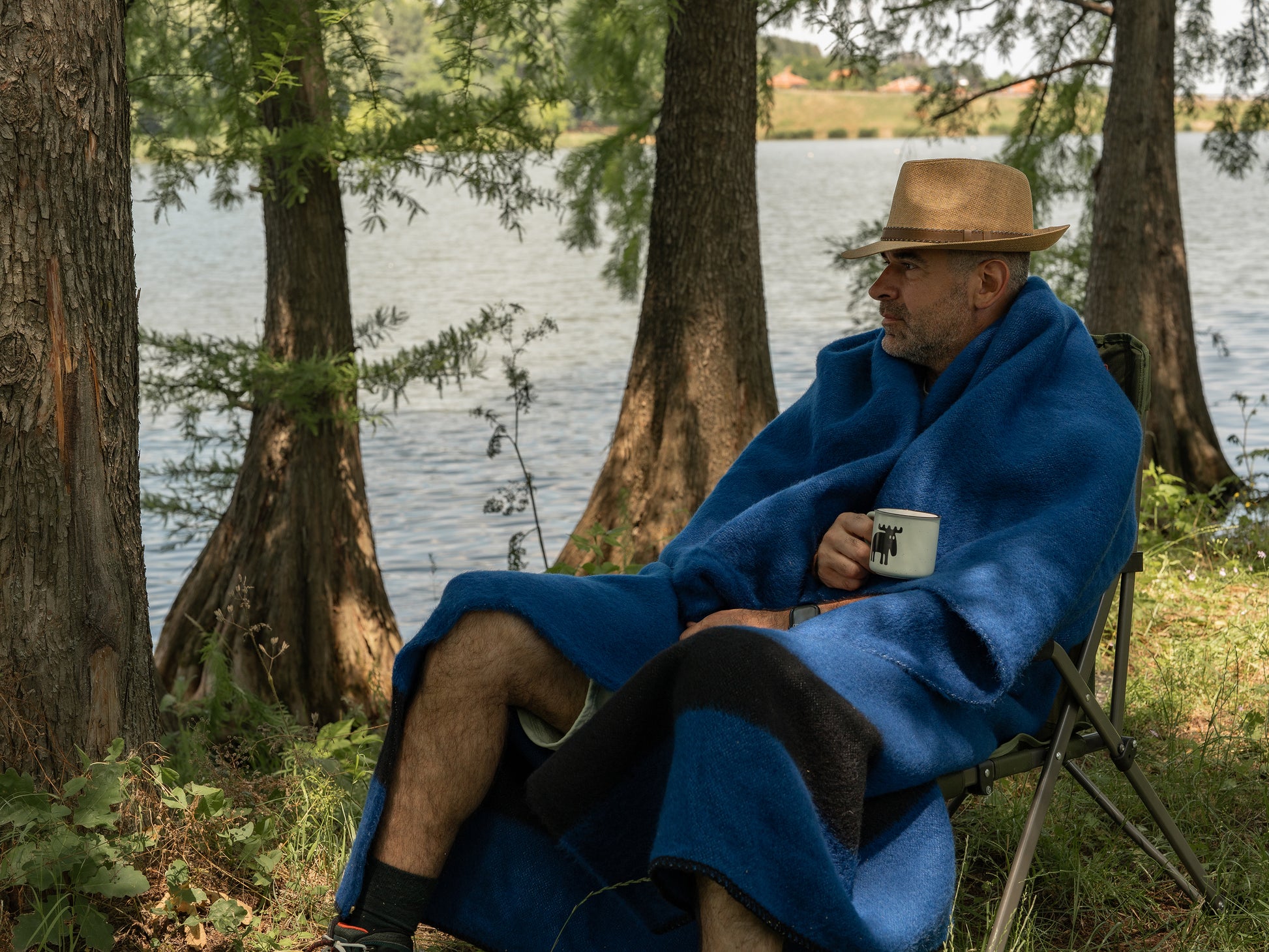 Person wrapped in a blue pioneer blanket with a black stripe, made of natural wool. The man is sitting in a chair by a lake, holding a mug.