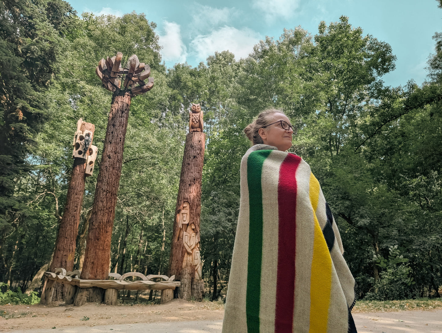 Woman wrapped in a heirloom iconic multi striped blanket standing in the forest with a wooden sculpture behind.