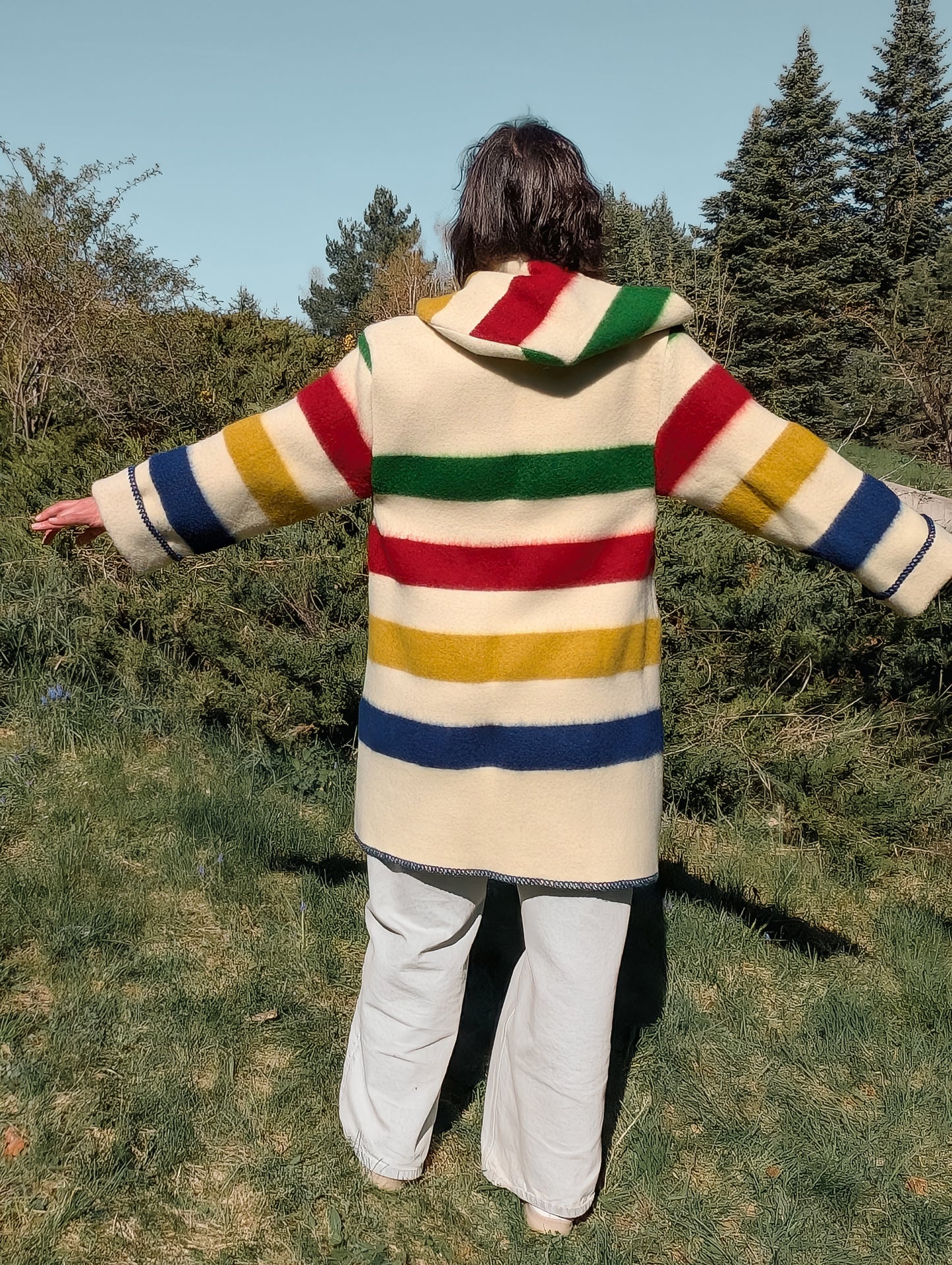 Woman wearing colorful striped overcoat with hood, combined with wide leg jeans, out in a park setting.