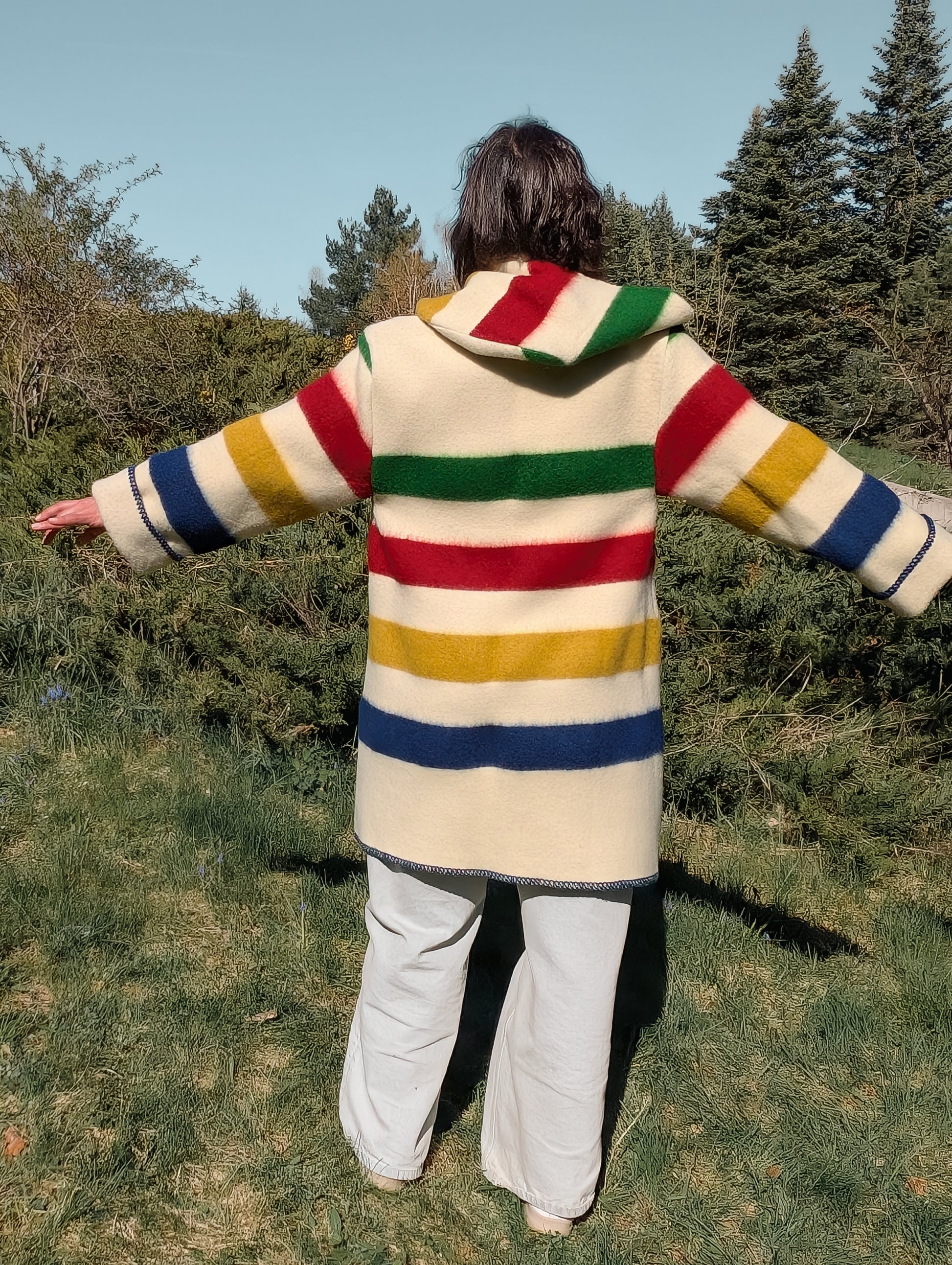 Woman wearing colorful striped overcoat with hood, combined with wide leg jeans, out in a park setting.
