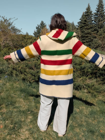 Woman wearing colorful striped overcoat with hood, combined with wide leg jeans, out in a park setting.