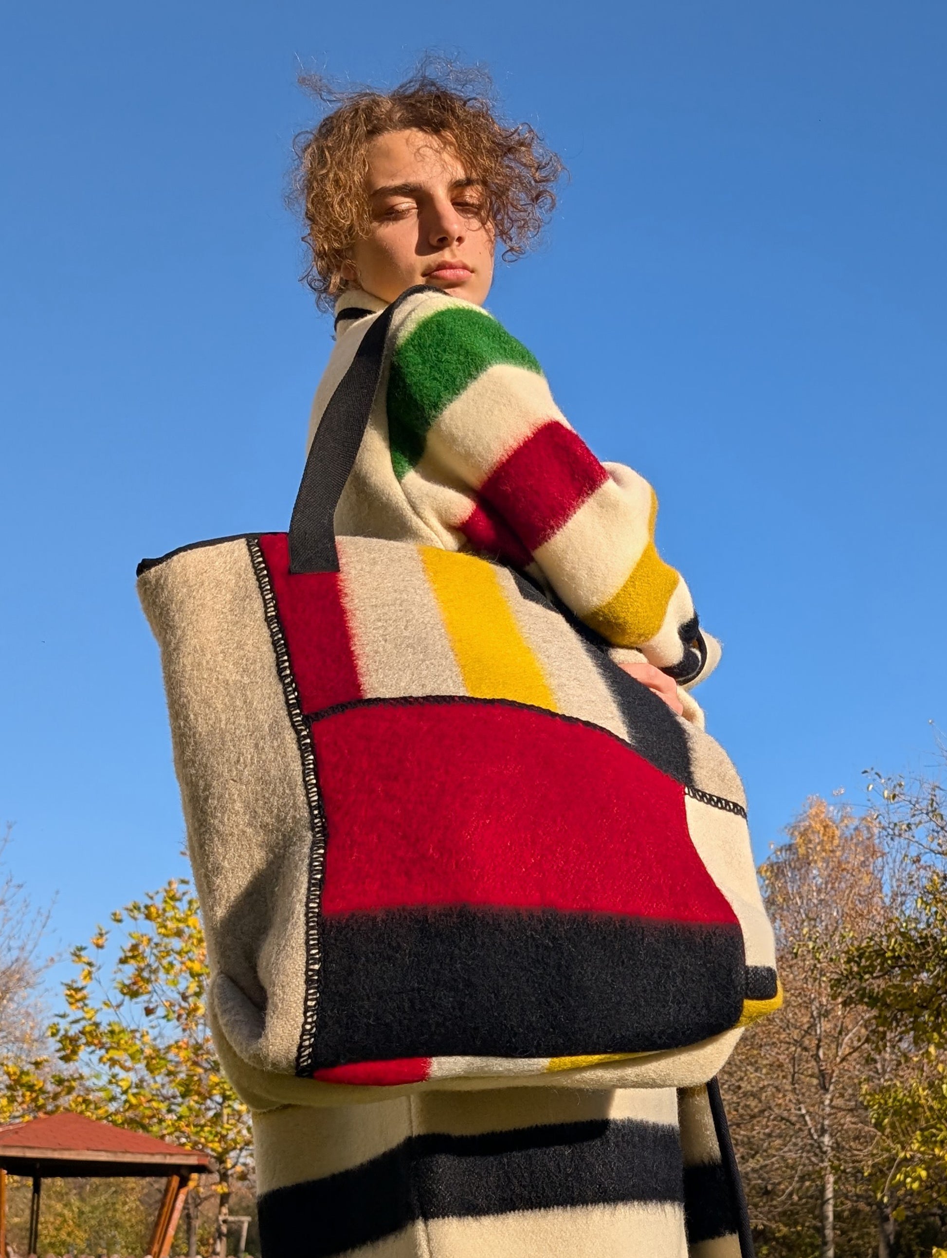 A young man wearing a large designer patchwork tote bag on his shoulder, matching his upcycled stripe duster coat. 