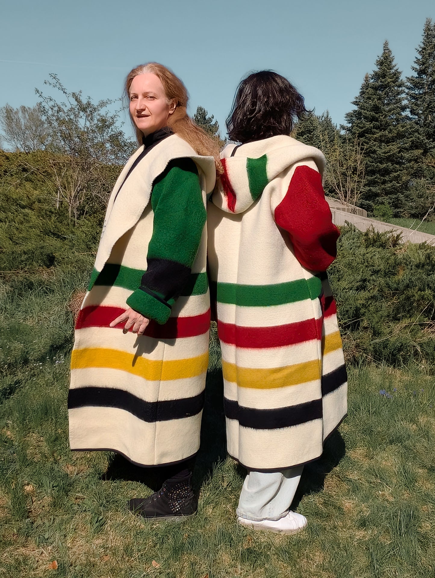 Two people wearing loose unisex woolen blanket overcoats with iconic colorful stripes and large hoods.