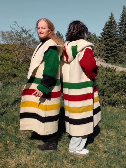 Two people wearing loose unisex woolen blanket overcoats with iconic colorful stripes and large hoods.