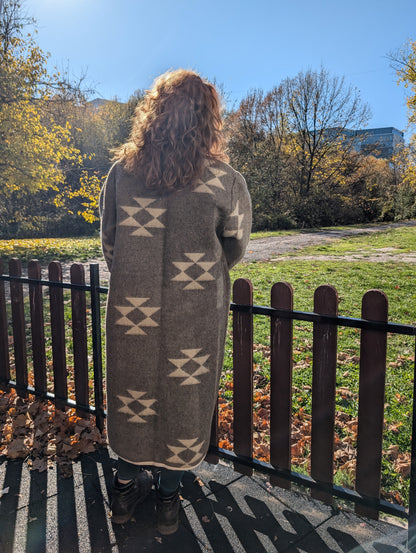 A woman standing backwards near a fence and wearing a tailored long wool cardigan coat made from undyed handmade wool blanket with aztec diamond symbols