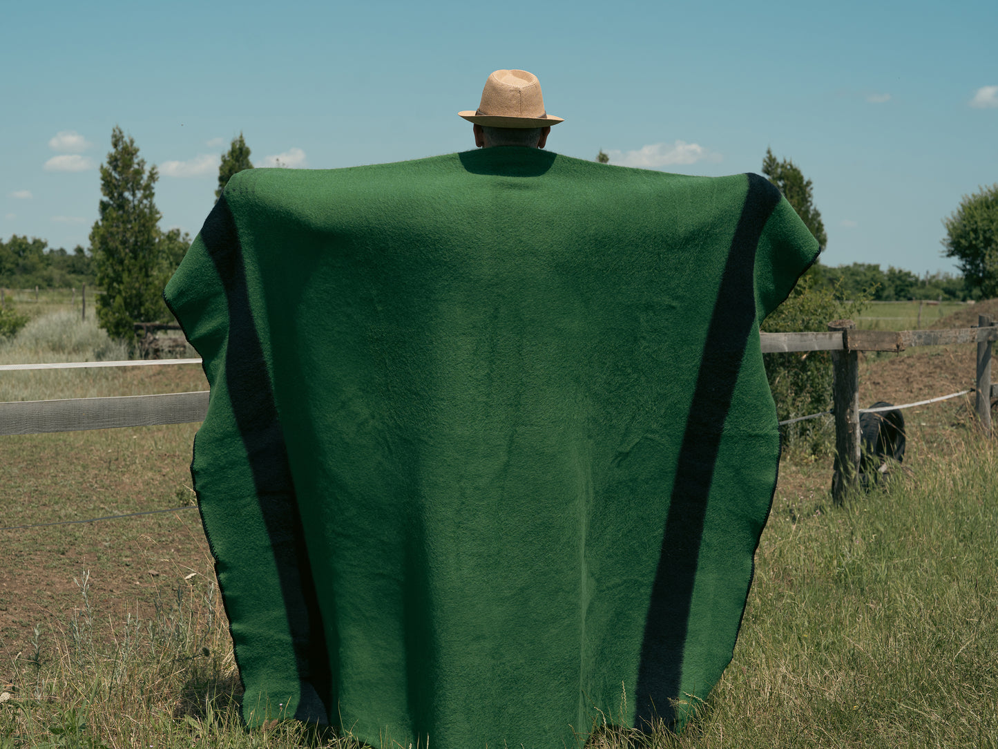 Person with hat covered with a green camp blanket standing in a field with a clear sky.