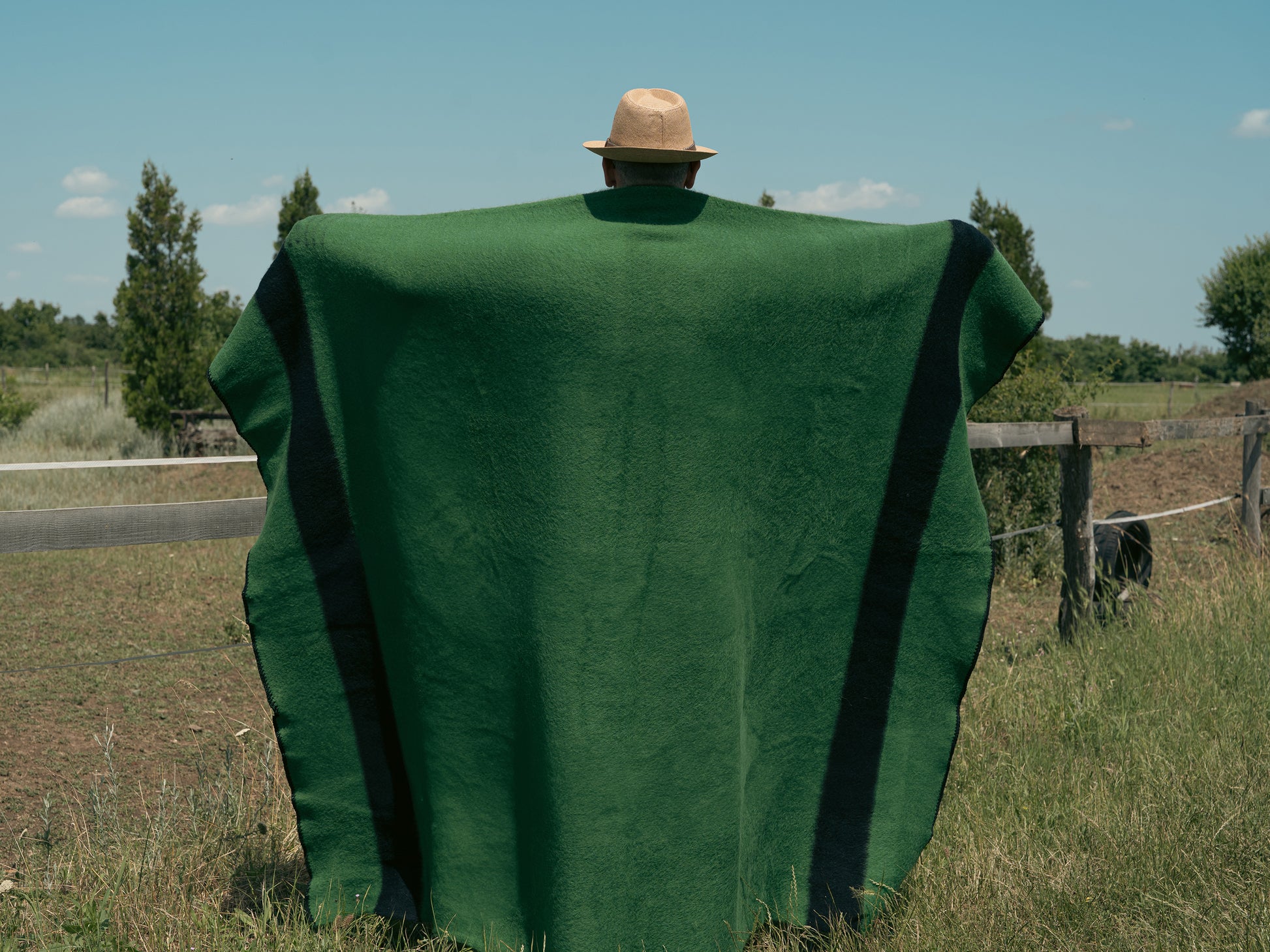 Person with hat covered with a green camp blanket standing in a field with a clear sky.