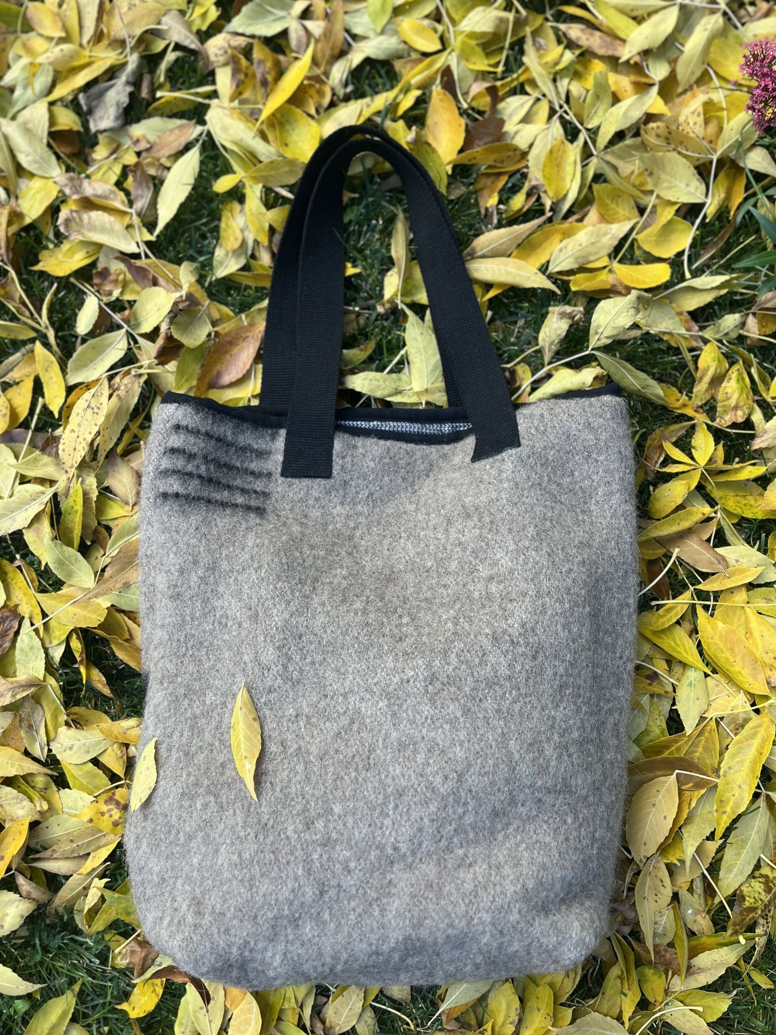 The back side of a gray wool blanket tote bag with black handles on a bed of yellow leaves.