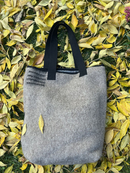 The back side of a gray wool blanket tote bag with black handles on a bed of yellow leaves.
