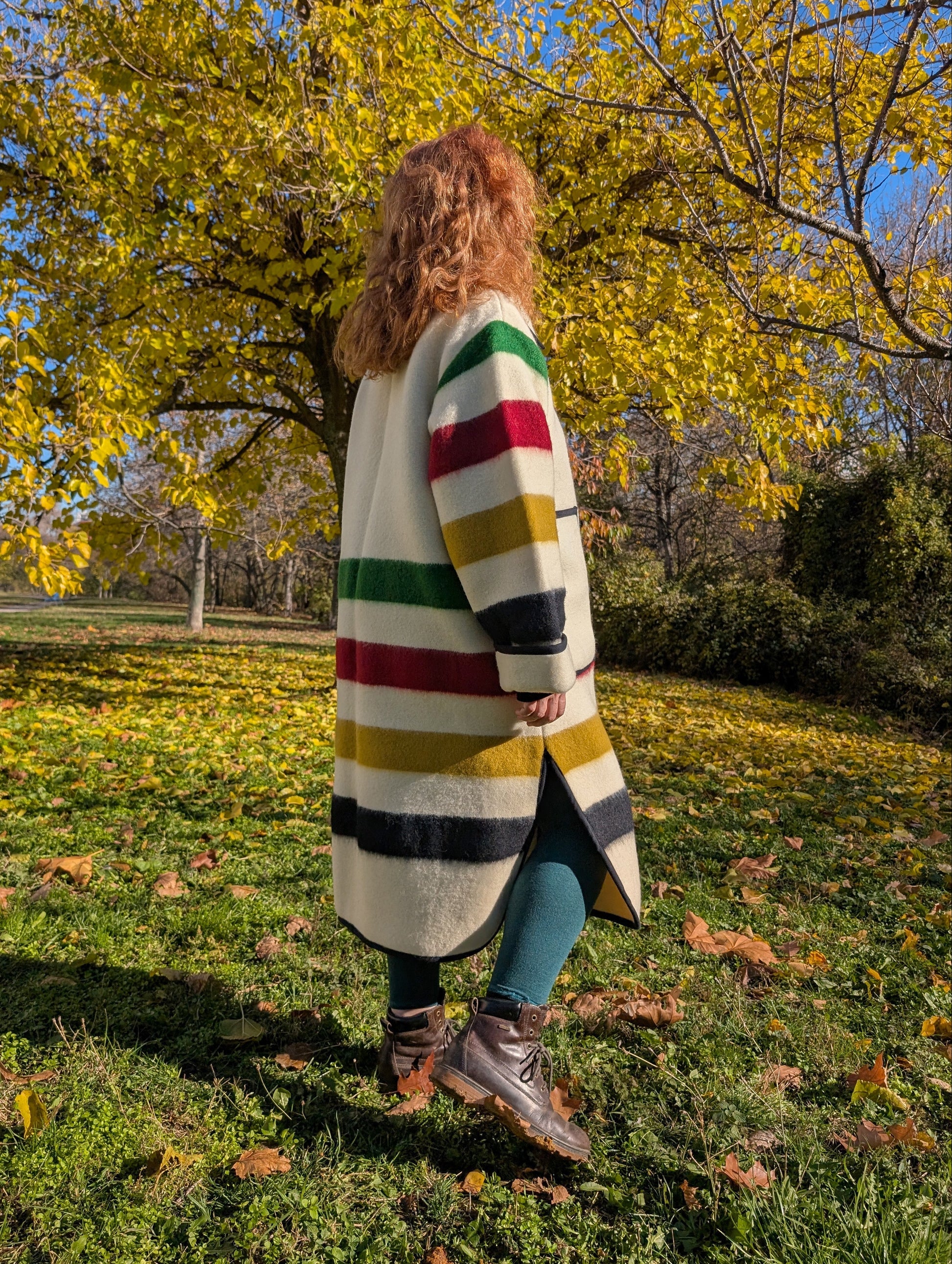 A woman posing at one side with a duster coat featuring side slits, classic collar and button closure. The multi stripes may refer to the Canada history and the fur trade.