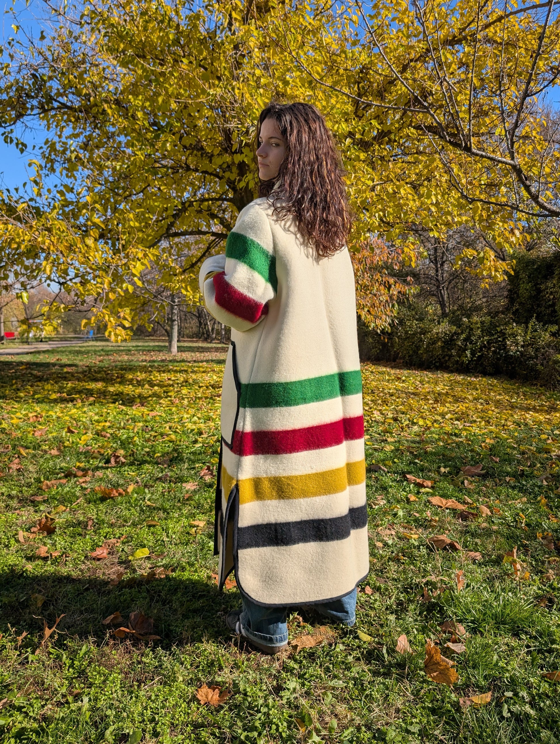 Person wearing a colorful striped coat standing in a park with trees and grass. Offwhite duster coat with stand collar, upcycled from a multistripe blanket. Oversized coat in cream base. Thick wool coat, single breasted coat.