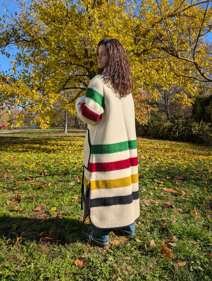 Person wearing a colorful striped coat standing in a park with trees and grass. Offwhite duster coat with stand collar, upcycled from a multistripe blanket. Oversized coat in cream base. Thick wool coat, single breasted coat.
