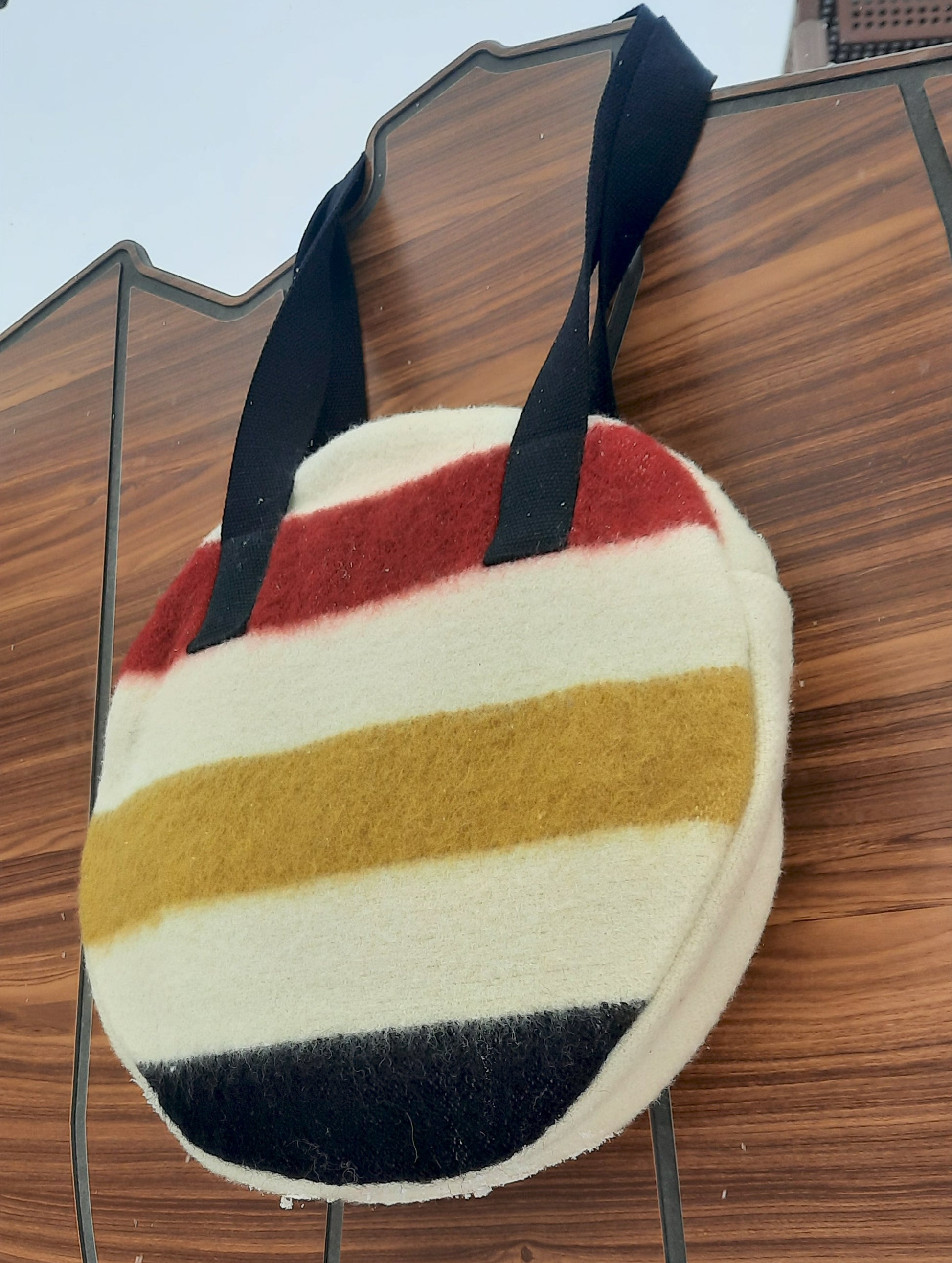 Round multicolor striped woolen blanket purse with black straps on a wooden surface