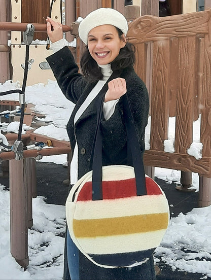 Woman holding a multistripe round tote bag with black straps. Handmade upcycled from pure wool frontier blanket.