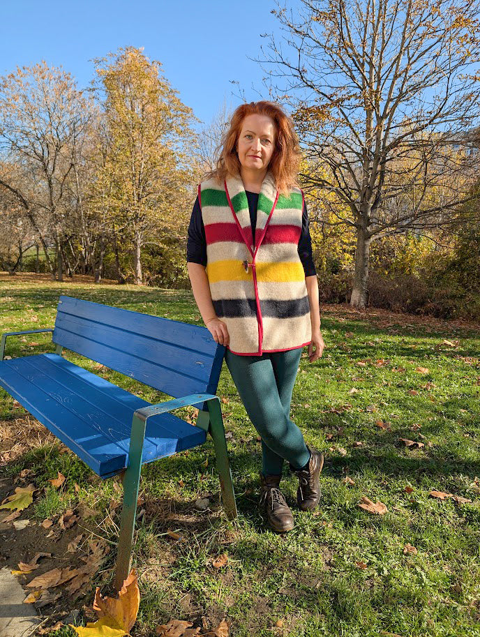 Woman wearing a multistripe wool blanket vest standing next to a blue bench in a park with trees and grass.