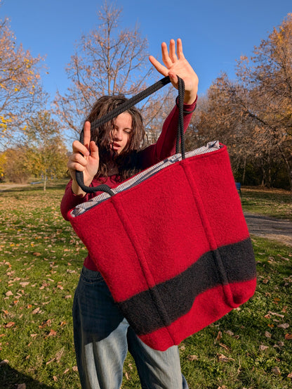 A young woman showing a large stylish red shopper bag with black robe straps and a stylish black stripe, upcycled from pure wool blanket.