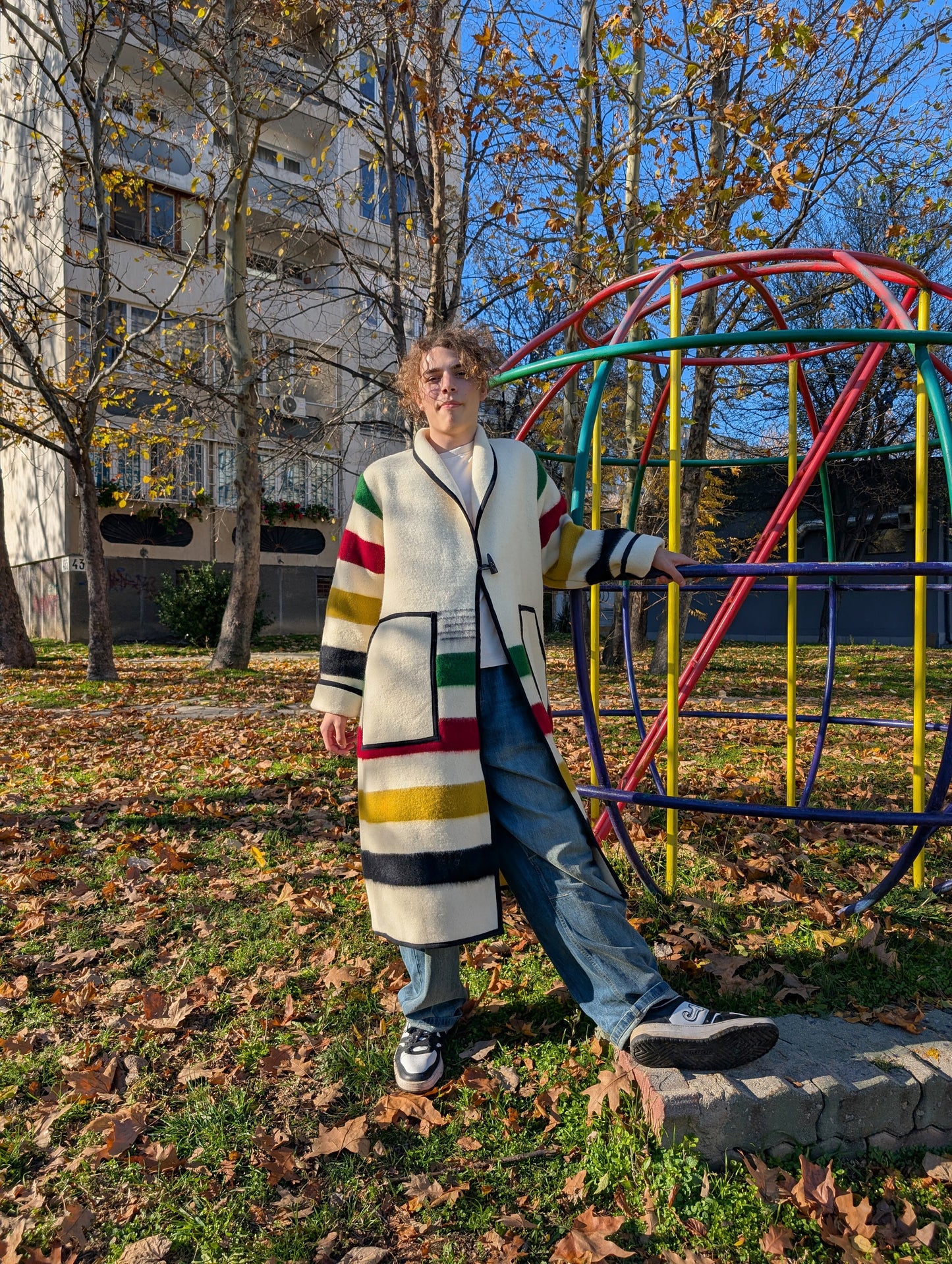 A young man wearing a loose long handmade robe coat, upcycled from a pure wool  blanket with cream base and multi stripes. The man is leaning on a Jungle gym out in the park. 