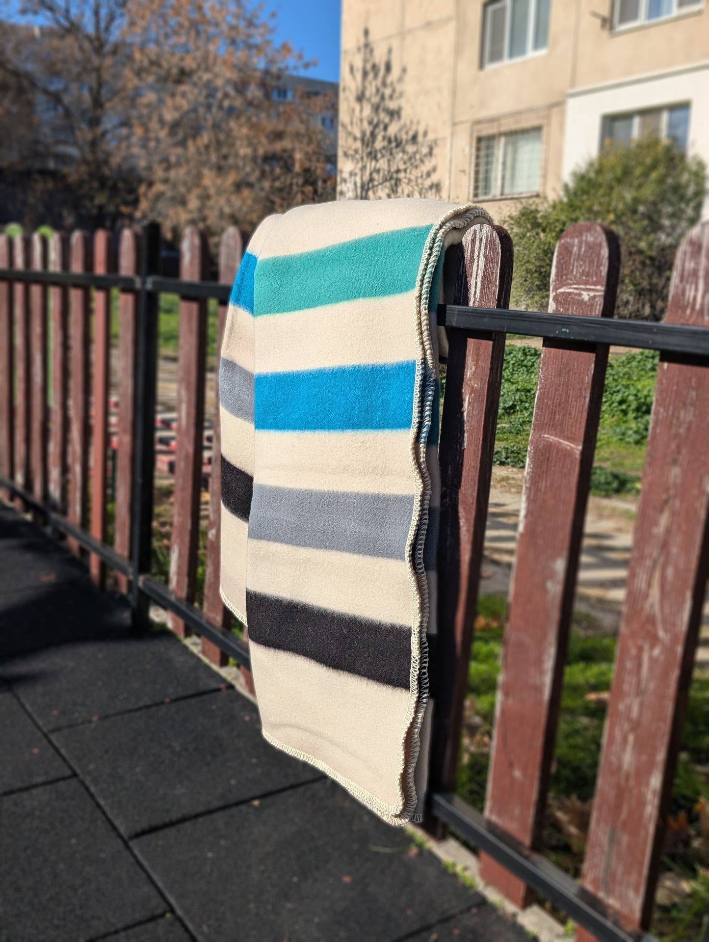 Handwoven striped blanket  in cream base with blue, gray, and black colors draped over a railing outdoors.