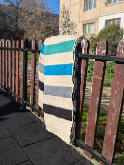 Handwoven striped blanket  in cream base with blue, gray, and black colors draped over a railing outdoors.