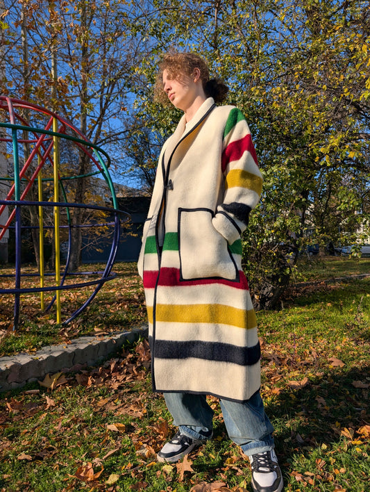 A young man posing with a long  off white shawl collar coat with toggle closure and two front pockets, tailored from sustainable wool blanket with traditional inspired stripes