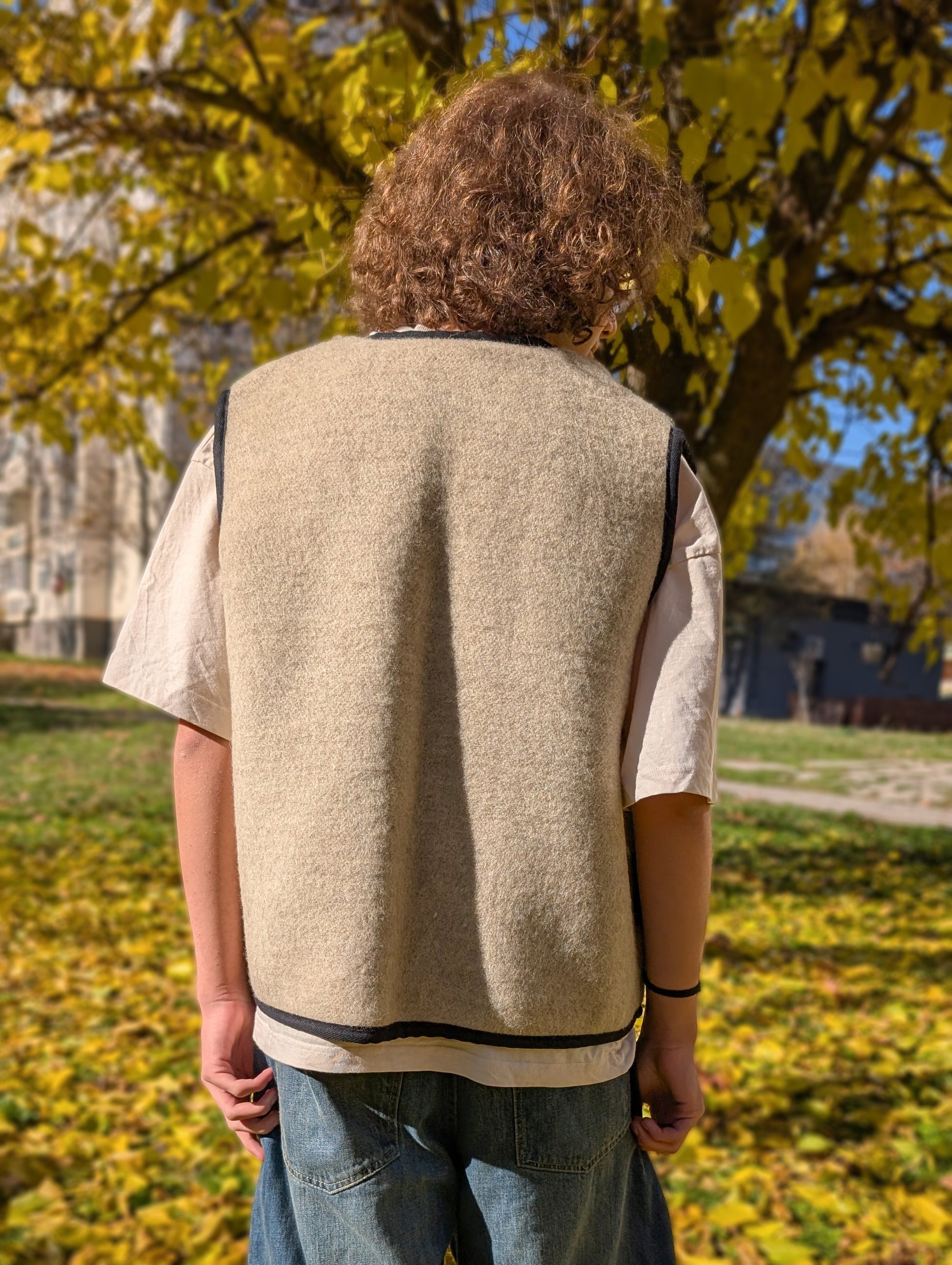 Man stading with his back, wearing a colorful stripe vest whose back is plain, the base color is earth gray or beige. The vest is upcycled from a pure wool blanket.