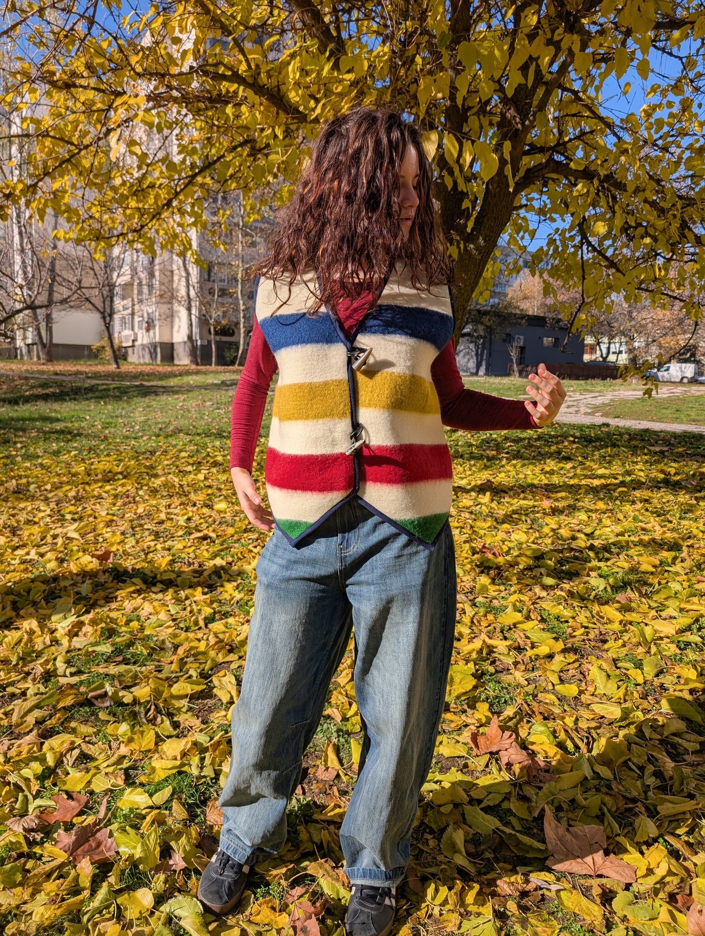 Person wearing a colorful striped sleeveless sweater in a park with autumn leaves. A made-to-order garment for slow fashion statement or premium quality wool.