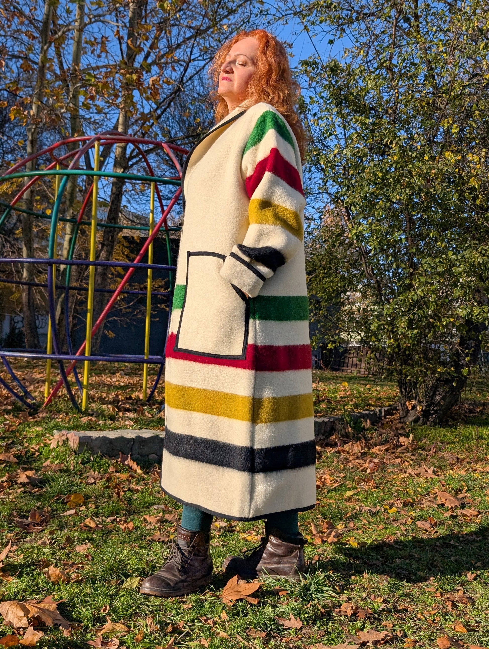 A woman wearing a loose long upcycled robe coat with shawl collar. The woman is leaning on a Jungle gym out in the park
