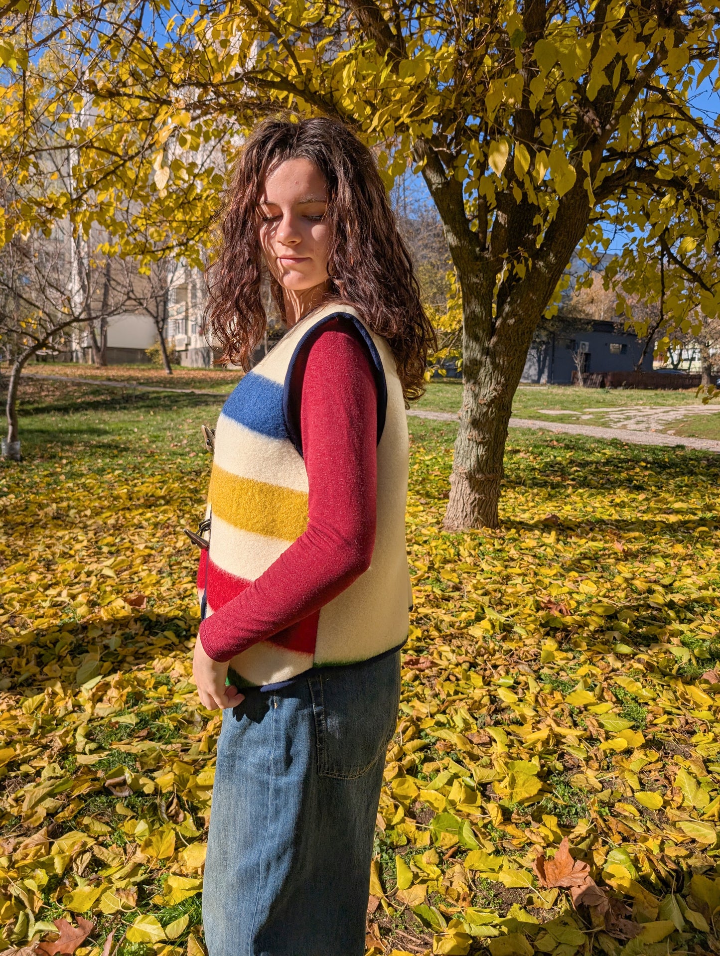 A girl wearing a unisex cropped all-purpose woolen vest with stripes in different colors at the front, and a plaini back. The base is ivory or off-white. 
