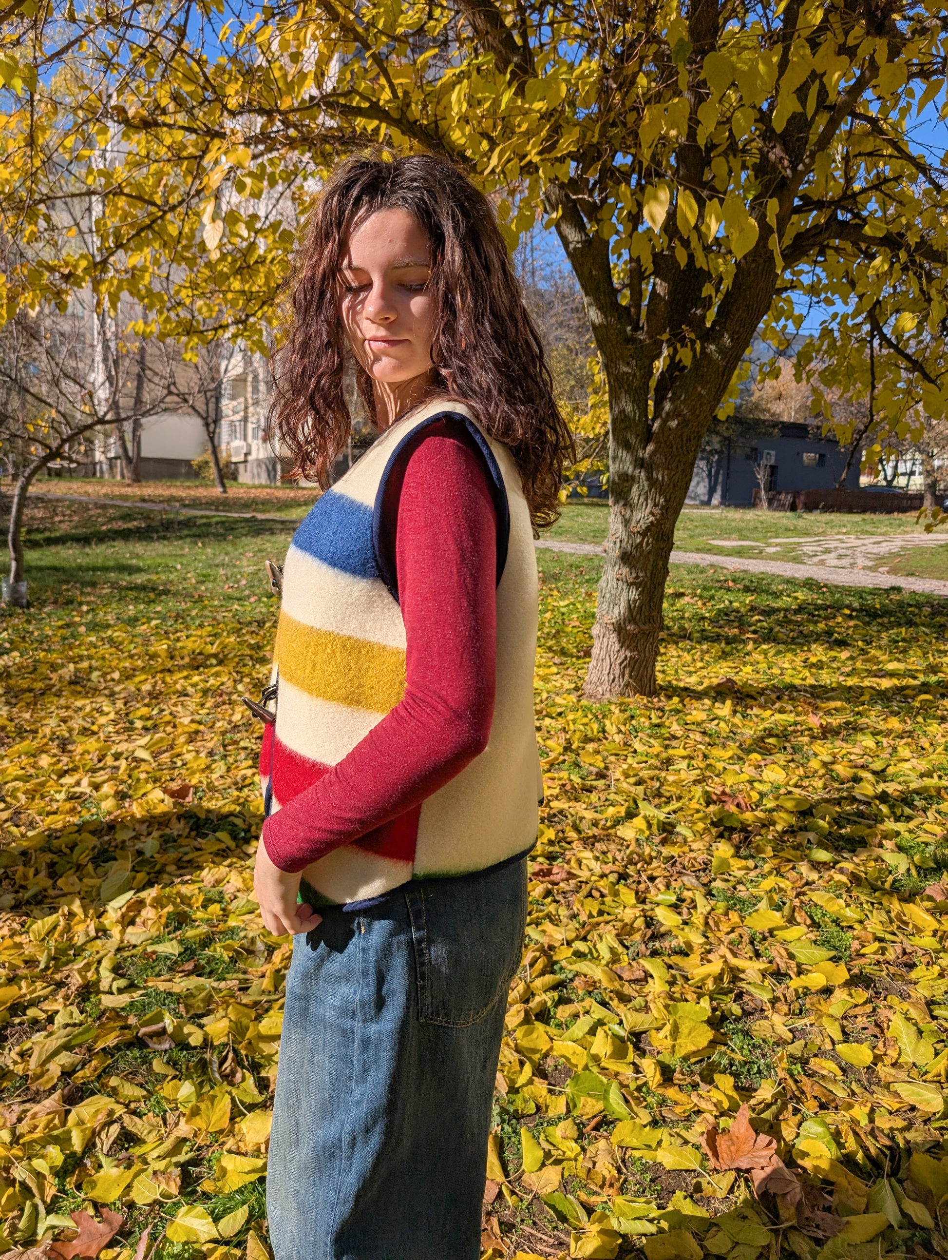 A girl wearing a unisex cropped all-purpose woolen vest with stripes in different colors at the front, and a plaini back. The base is ivory or off-white. 
