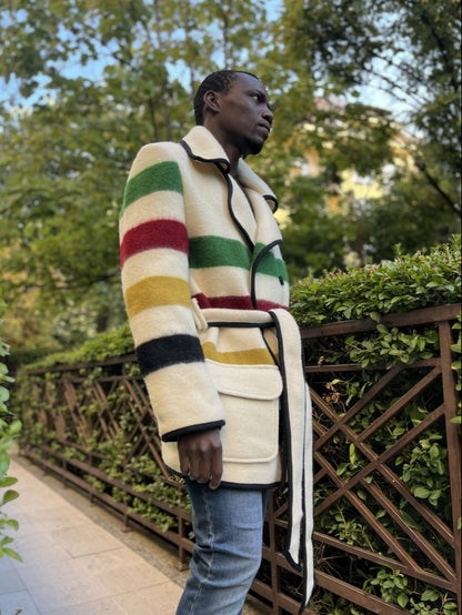 Person wearing a colorful striped blanket coat with double breasted closure, kinched with a belt in an outdoor setting with greenery.