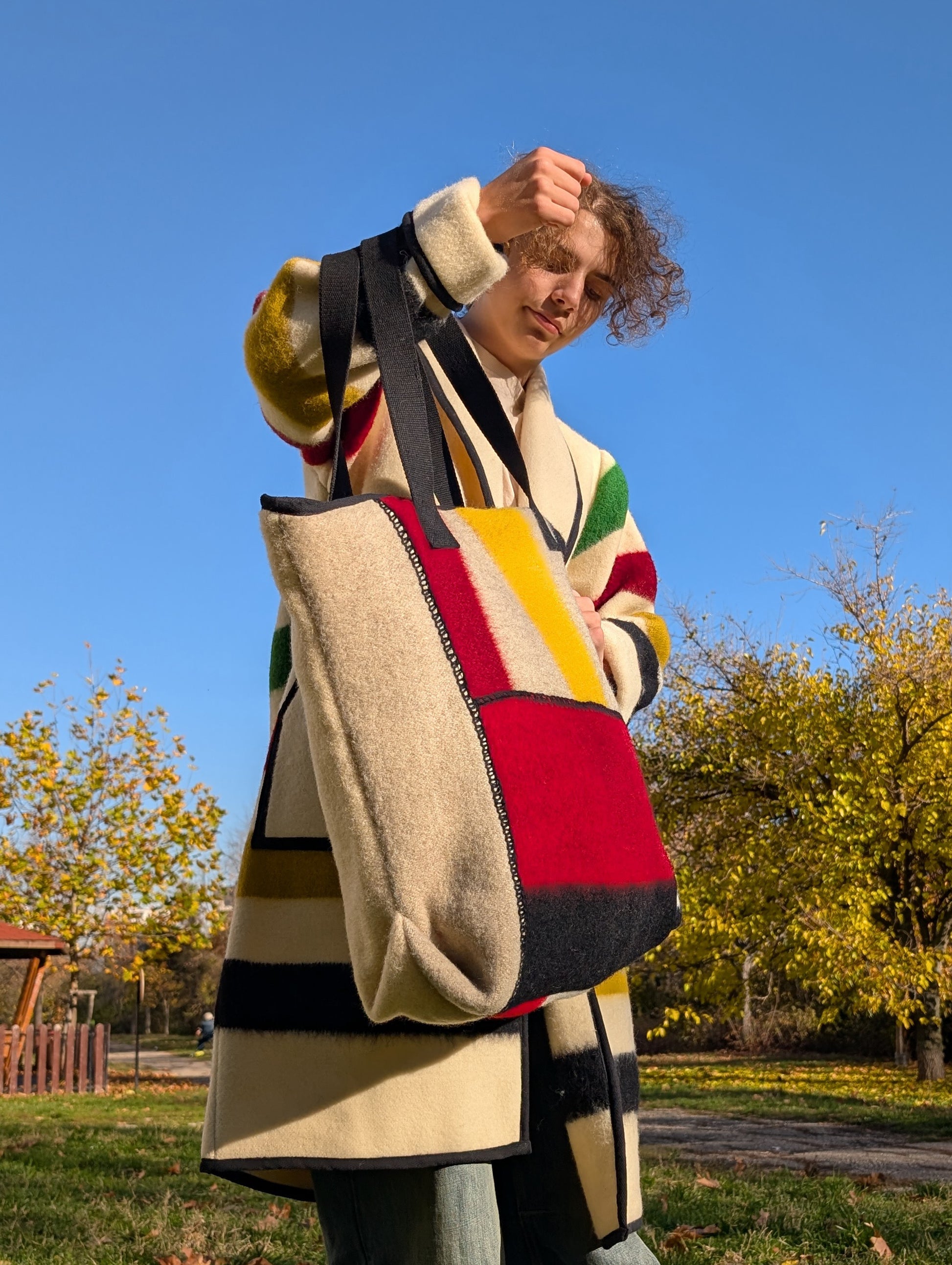 Person holding a colorful tote bag made from wool blanket patches and matching his upcycled stripe robe coat. Both are a slow fashion statement.