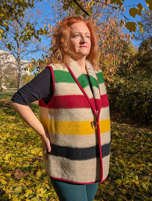 Woman wearing a stylish woolen  blanket vest with multicolor stripes, a shawl collar and a single toggle horn closure. The stripes run horisontally along the front and the back. 