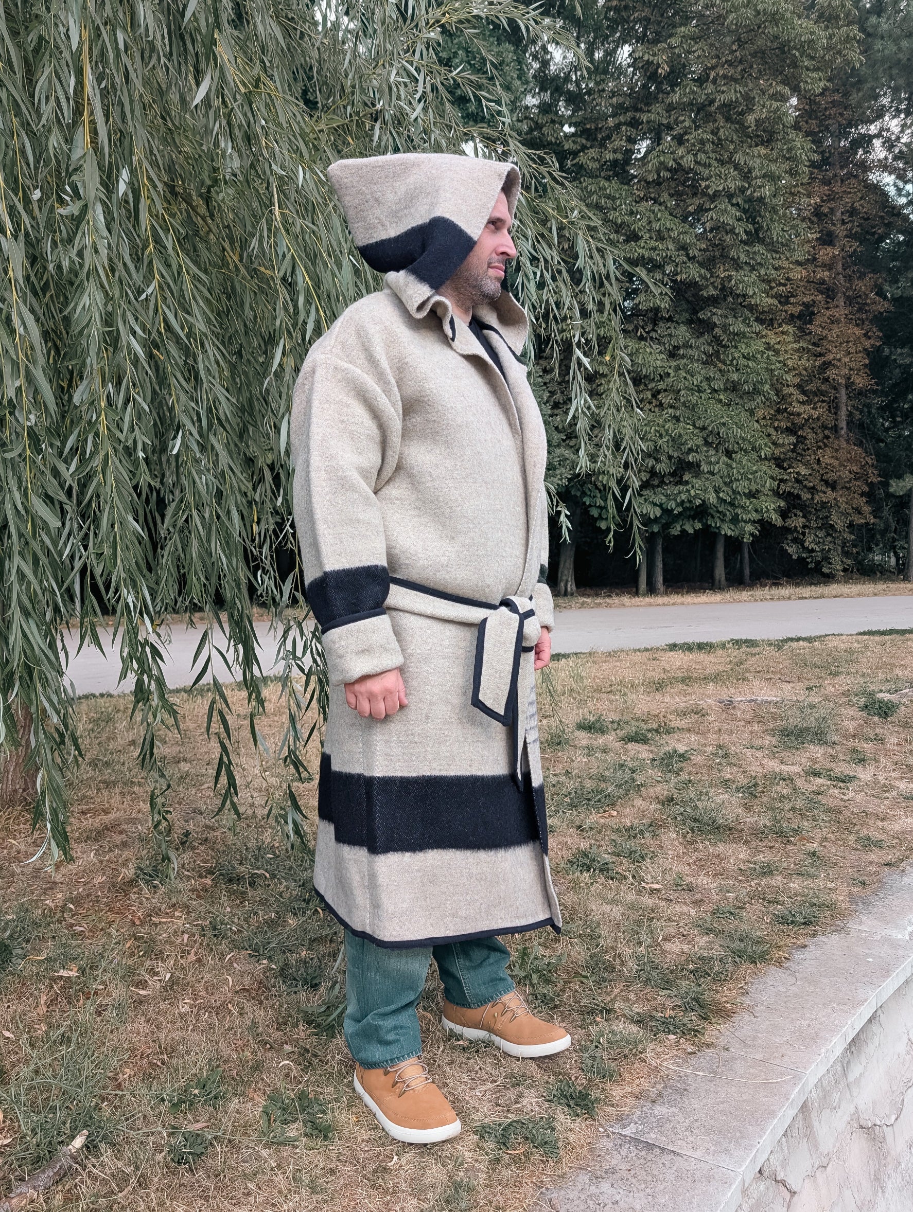 A man wearing an oatmeal wool blanket capote coat with a black stripe and large hood on,  made from repurposed traditional wool blanket.