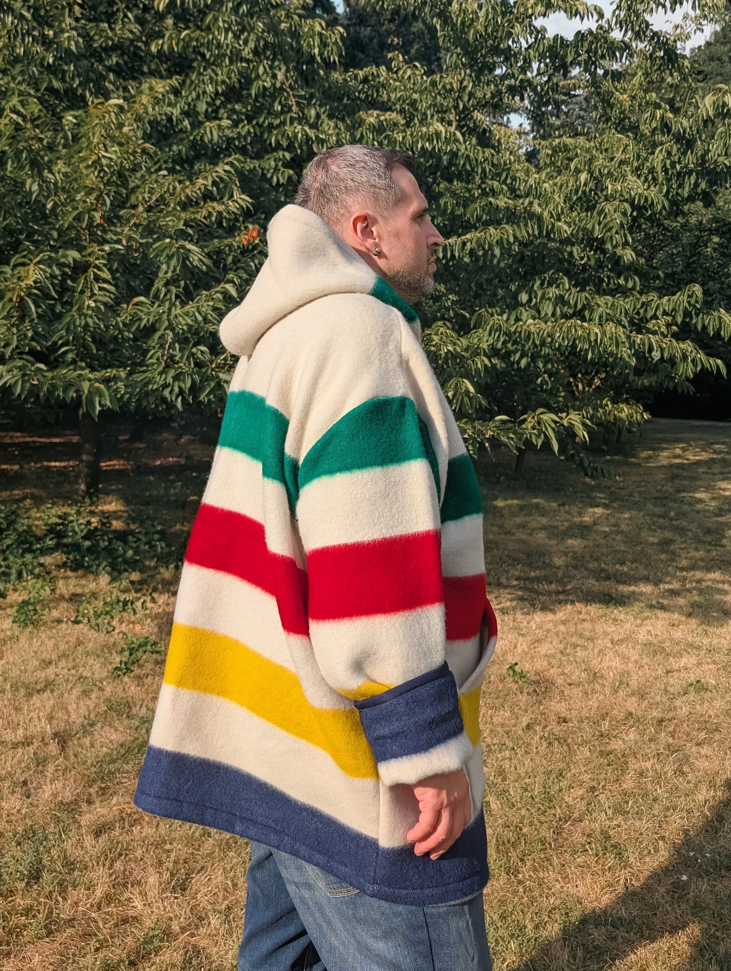 Man wearing an iconic large multistriped camp hoodie with ivory base and colorful stripes, with a large hood and a front cangaroo pocket. 