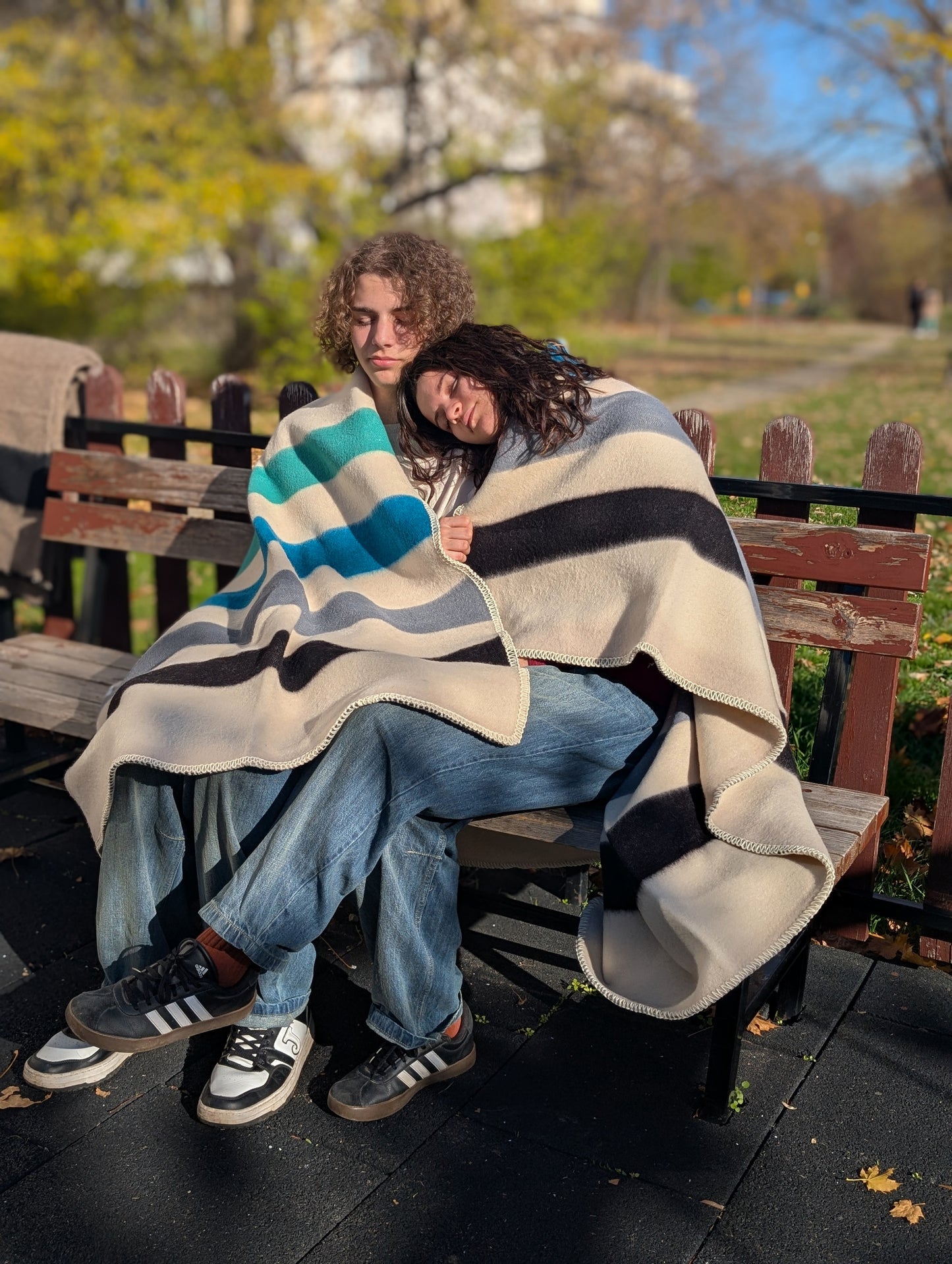Two people sitting on a bench under a premium quality slow processed multi color blanket with a park setting in the background.