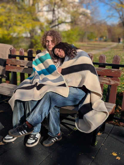 Two people sitting on a bench under a premium quality slow processed multi color blanket with a park setting in the background.