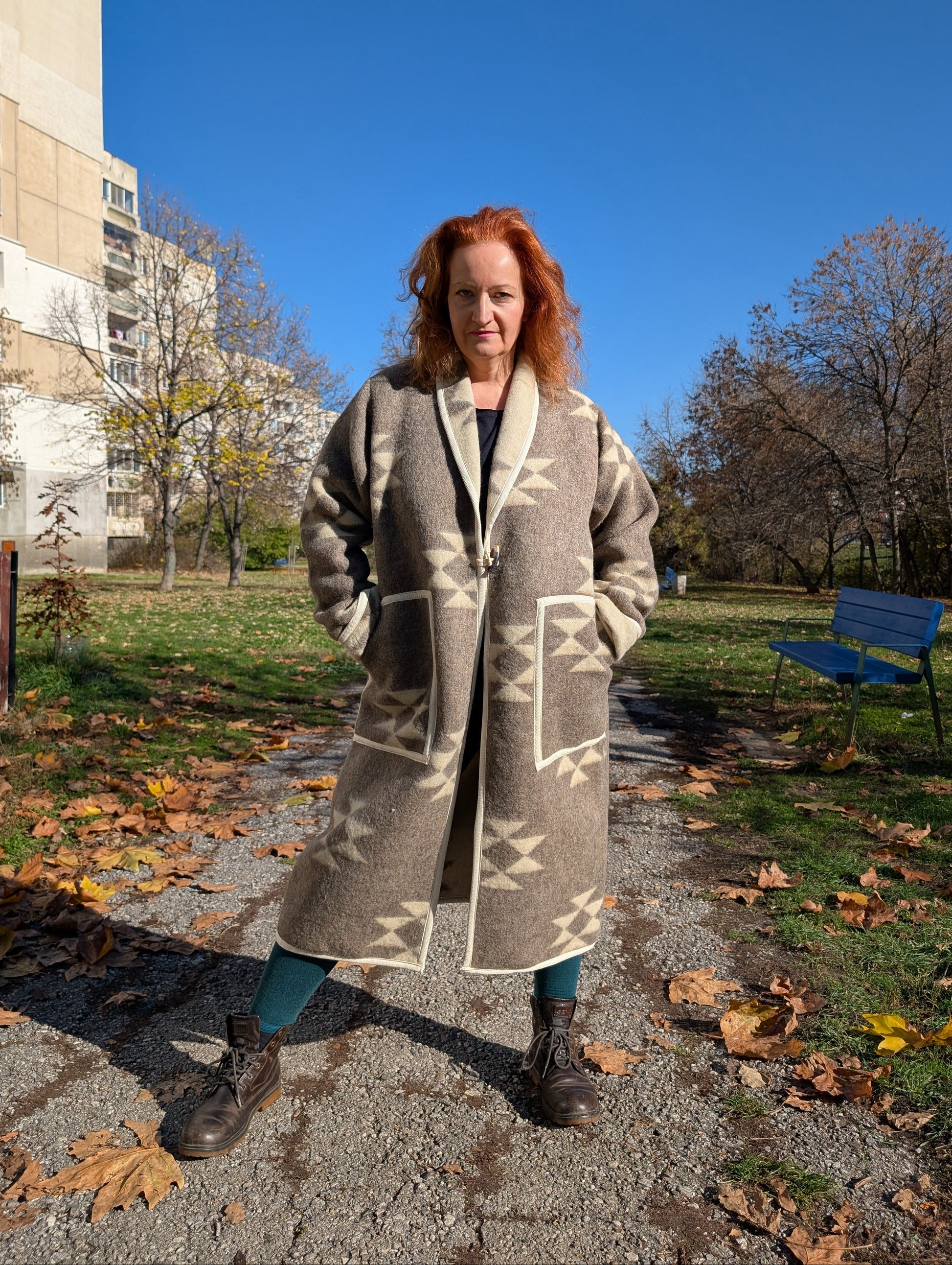 Woman wearing a long robe coat upcycled from undyed pure wool blanket with folk motifs. standing on a pathway outside.