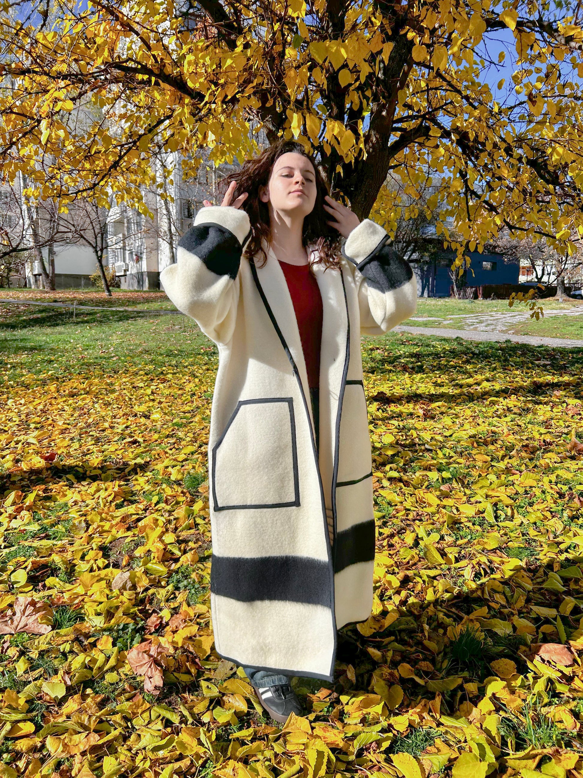 A woman wearing a loose off-white robe coat tailored from a pure virgin wool blanket with black stripes. The coat features a robe collar, two front pockets and a toggle closure. 