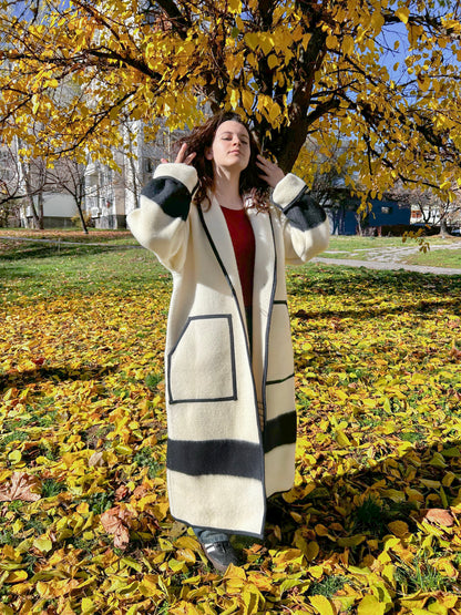 A woman wearing a loose off-white robe coat tailored from a pure virgin wool blanket with black stripes. The coat features a robe collar, two front pockets and a toggle closure. 