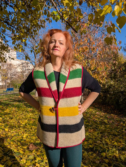 Woman wearing a cool blanket gilet in earth gray or oatmeal base with multistripe design and a single horn toggle. A bespoke garment, tailored from a repurposed handmade wool blanket.