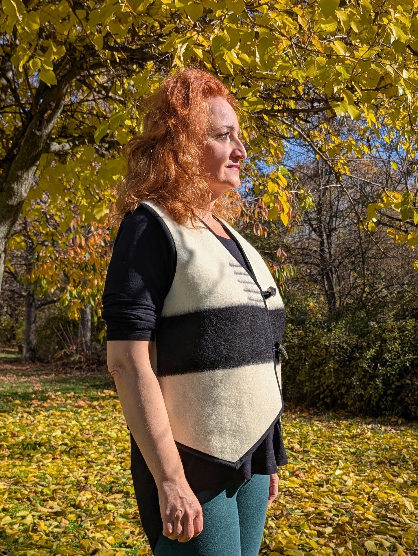 Woman wearing a classic woolen vest with V-ront, in cream white with a black stripe running horisontally across the front, and toggle closure. 